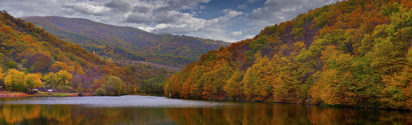 A wide valley landscape with a lake surrounded by hills covered in colorful autumn foliage under a partly cloudy sky, scenic and reflective mood.